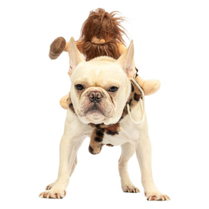 A cream-colored French Bulldog wearing a 'Paleolithic Woman' costume, featuring a brown spotted leopard-print skirt, a rope harness, and a brown faux-fur wig with a doll attached on its back, photographed against a solid white background.