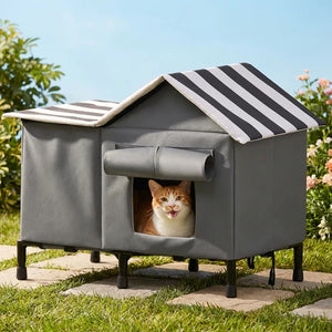 A gray outdoor cat house with a black-and-white striped roof, shown on a stone path in a garden with a cat peeking out from the front window at Pet Servo