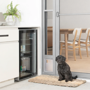 Dog sitting on a mat in front of an open refrigerator in a modern kitchen.
