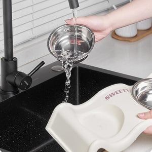 A stainless-steel pet water bowl being filled with running water from a black faucet, held over a black kitchen sink. A cream-colored pet feeder or scoop with a small stainless-steel bowl is also visible in the foreground, with a hand holding it at Pet Servo