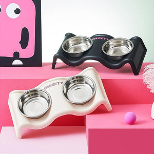 Two pet feeding stations with stainless steel bowls, one cream and one black, displayed on pink geometric blocks with a pink cartoon card in the background at Pet Servo