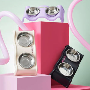 Three pet double bowls with silicone holders in lavender, cream, and black, displayed on pink pedestals against a mint green background at Pet Servo