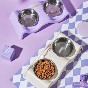 Two pet feeding stations arranged on a lavender and white checkered surface. The top station is lavender with stainless steel bowls, and the bottom station is cream with one bowl filled with kibble at Pet Servo