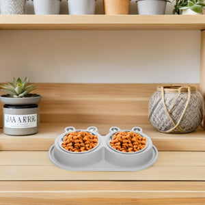 A gray silicone double pet food bowl with two small, raised bowl edges, shown on a wooden shelf with food inside. The scene includes home decor props like a small potted succulent and a woven basket at Pet Servo