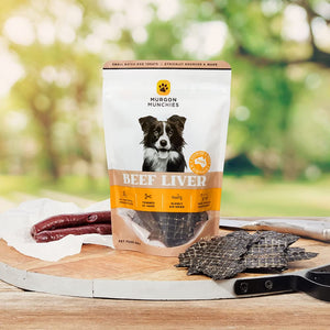 A stand-up pouch of Murgon Munchies Beef Liver dog treats stands on a wooden cutting board outdoors, with several dark cured meat strips and a few square jerky-style treats placed around it on a wooden table. The background is a softly blurred green outdoor setting.