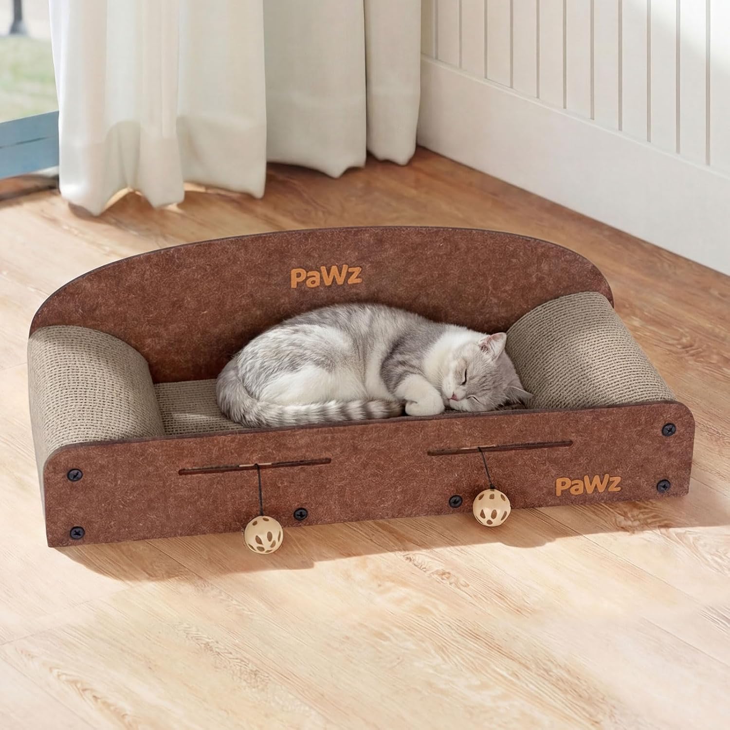A gray and white cat is curled up sleeping inside a brown and beige cat bed with dangling toy balls. The cat bed has the brand name 'PaWz' printed on the front, set on a light wood floor in a room with white curtains at Pet Servo