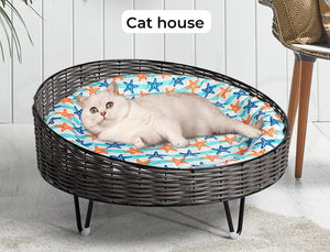 A white cat lies inside a round woven pet bed with a blue, orange, and white starfish-patterned cushion. The bed has a dark gray wicker frame and small black legs, set against a white paneled wall with a chair partially visible on the right at Pet Servo