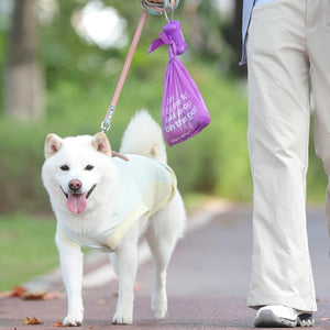 White dog on a leash with a purple bag attached, walking on a path in a park at Pet Servo