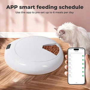 A white round smart pet feeder sits on a wooden floor, with a white cat standing nearby and a hand holding a smartphone app screen. Text at the top reads 'APP smart feeding schedule' and says the app can pre-set up to 6 meals per day at Pet Servo