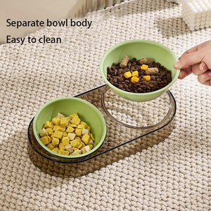 A two-piece pet food bowl set with light green bowls on a clear acrylic stand, shown on a textured cream rug. One bowl contains dry kibble and the other contains wet cubes, with a human hand holding the top bowl at Pet Servo
