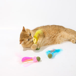 A golden cat lies on a white studio floor while playing with a yellow feather toy. Two additional colorful feather and ball toys are placed nearby on the floor at Pet Servo