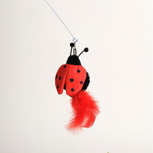 A red and black ladybug-shaped hanging toy with black antennae and a bright red feather dangling below, photographed against a plain white background at Pet Servo