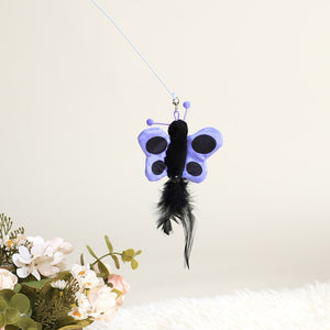 A purple and black butterfly-shaped cat toy with a black feather dangling below, suspended from a thin white string against a warm off-white background, with decorative flowers in the lower left corner at Pet Servo