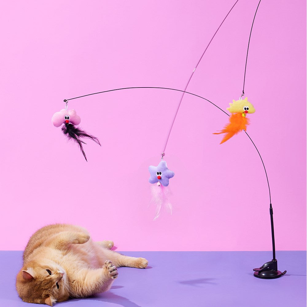 A ginger cat lies on its back on a purple surface, looking at a black cat toy with three dangling colorful plush toys against a pink and purple studio background at Pet Servo