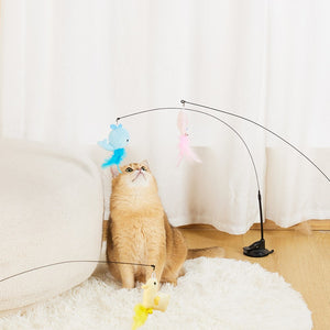 A fluffy orange cat sits on a white rug in a bright room, looking up at a black arched cat toy with dangling plush toys in blue, pink, and yellow at Pet Servo