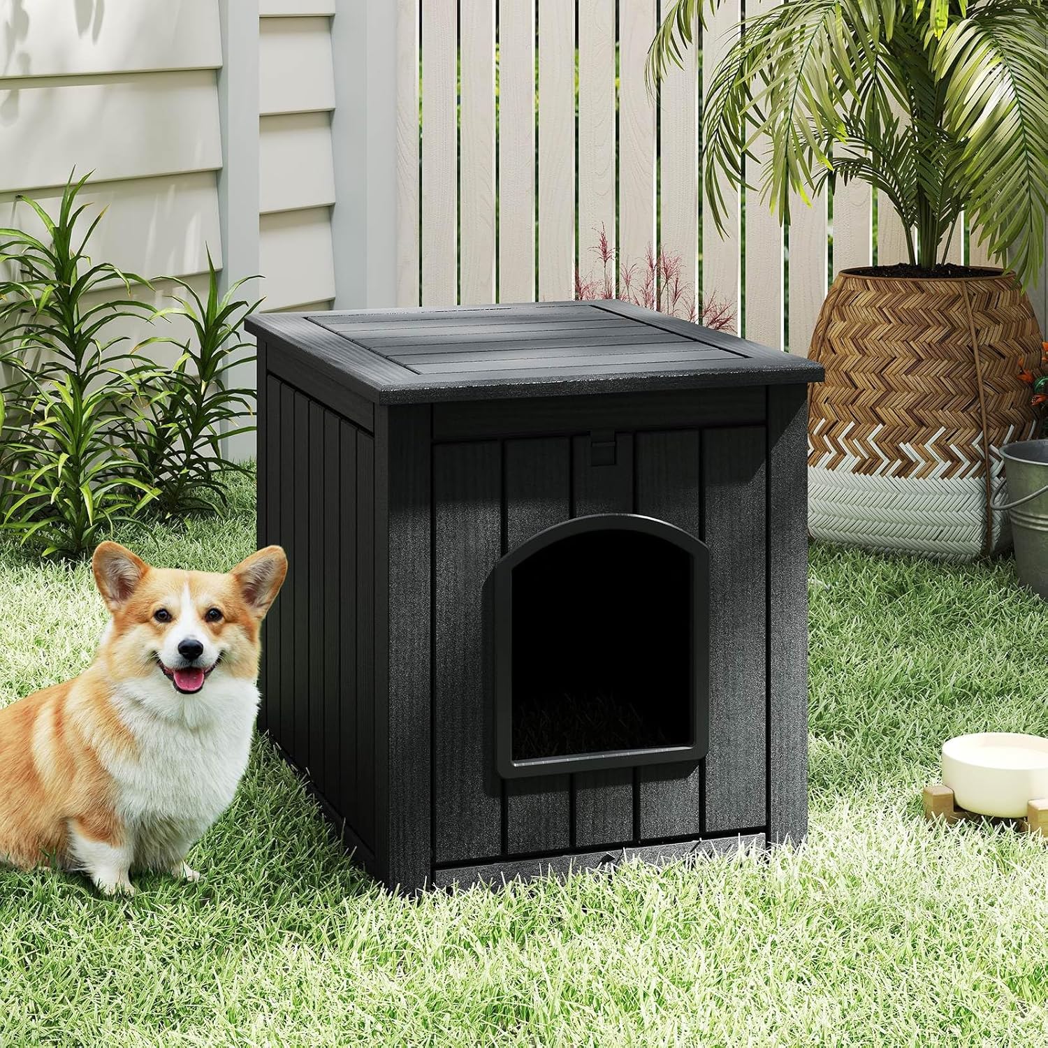 Dog sitting next to a black wooden doghouse on grass with plants and a house in the background at Pet Servo