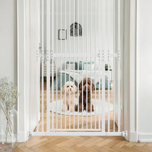 A white metal pet gate with vertical bars stands in an open doorway, separating two small dogs in a bright bedroom. The scene is styled with light wood flooring, white walls, and a vase of dried stems in the foreground at PET SERVO