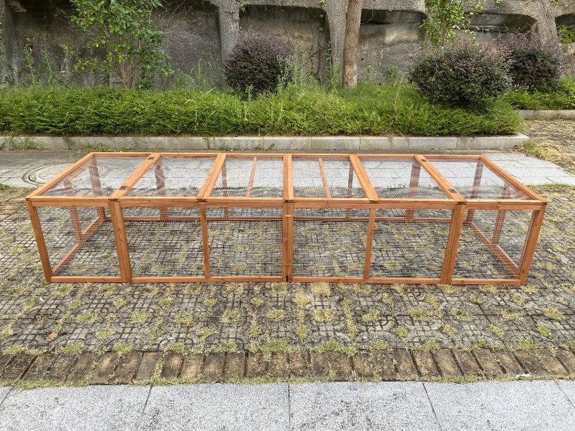 A long wooden frame structure with clear panels, photographed outdoors on a stone-paved surface with shrubs and a stone wall in the background at Pet Servo