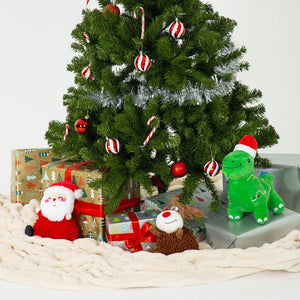 A decorated artificial Christmas tree with silver tinsel, red ornaments, candy canes, and wrapped gift boxes underneath, styled with plush toys including Santa Claus, a green dinosaur, and a reindeer on a cream knit blanket against a plain white background.