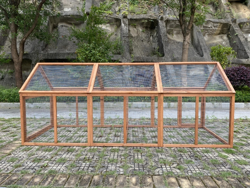 A long wooden chicken coop or small greenhouse with wire mesh panels, photographed outdoors on a paved surface in front of a rocky wall and trees at Pet Servo