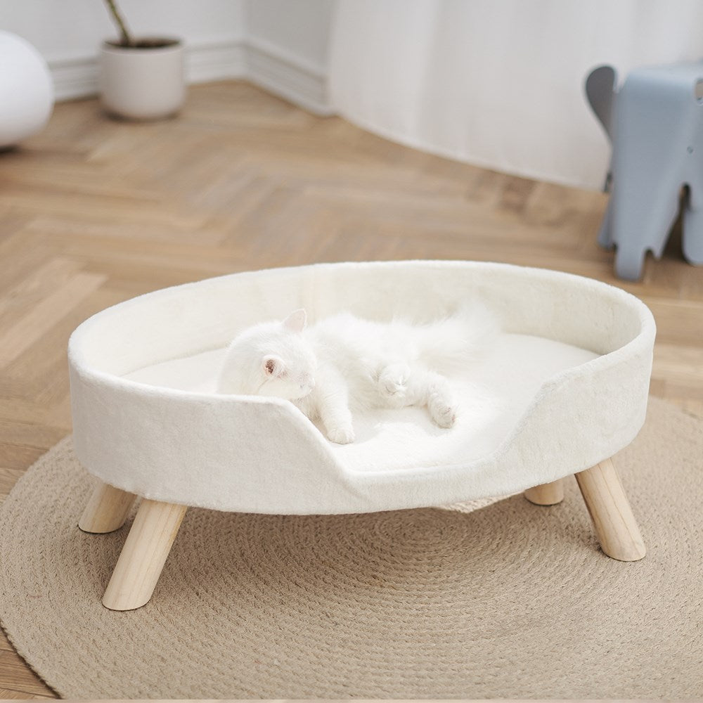 White pet bed with wooden legs in a room with a wooden floor and a plant in the corner at Pet Servo