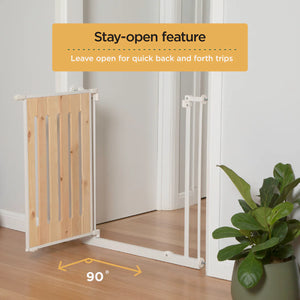 Wooden pet gate with stay-open feature labeled, on a wooden floor with a plant in the corner.