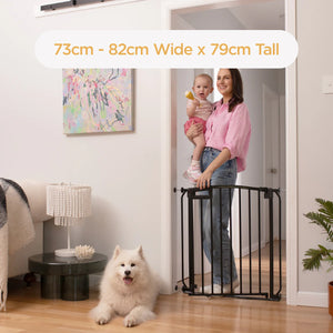 Woman holding a child and a dog standing behind a baby gate in a home setting.