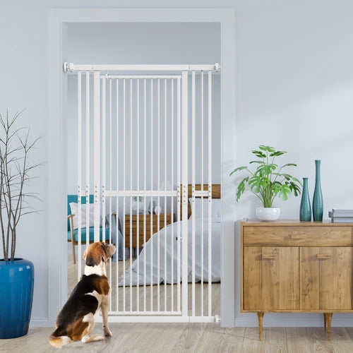 A white metal pet gate installed across a doorway in a bedroom, with a tricolor dog sitting in front of it on a light wood floor. The room includes a wooden cabinet, potted plants, and a bed in the background at PET SERVO