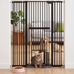 A black metal pet gate with a low-profile door and a small ramp, shown installed in a doorway in a home setting. A brown tabby cat is sitting on the ramp in the foreground, and a golden retriever dog is lying on the floor behind the gate at Pet Servo
