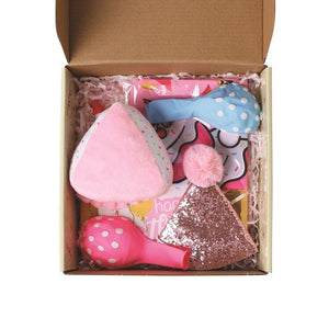 A top-down view of a brown cardboard gift box filled with pink and multicolor baby accessories, including a pink plush heart-shaped item, a blue polka-dot bow, a pink pom-pom, and a glittery pink triangle accessory, set against a plain white background at Pet Servo