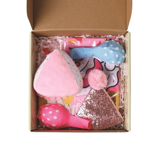 A top-down view of a brown cardboard gift box filled with pink and multicolor baby accessories, including a pink plush heart-shaped item, a blue polka-dot bow, a pink pom-pom, and a glittery pink triangle accessory, set against a plain white background at Pet Servo