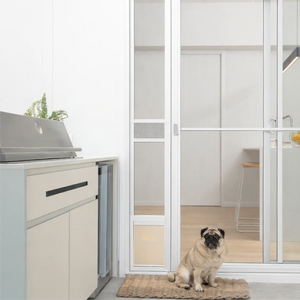 Pug sitting on a mat in front of a glass door with a view into a kitchen.