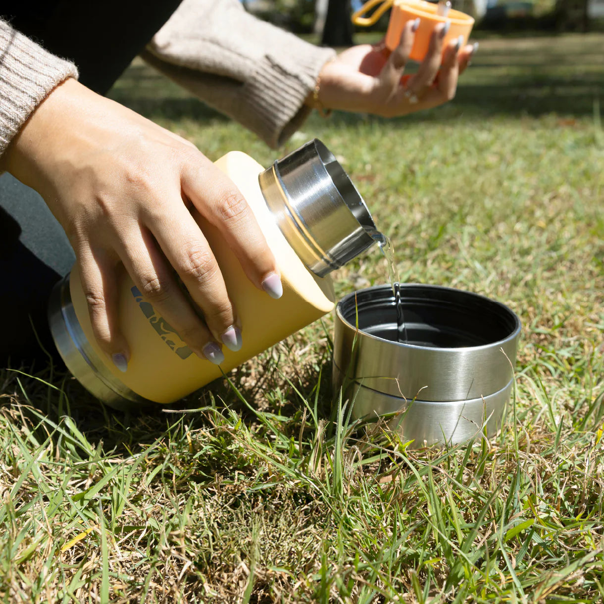 Person pouring liquid from a yellow thermos into a metal container on grass