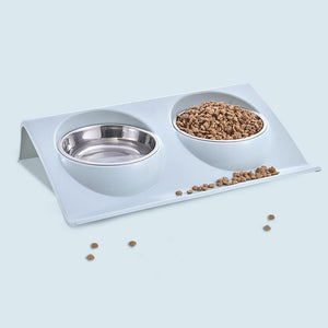 A white dual-bowl pet feeder and waterer stand with stainless steel bowls, shown on a light blue background with some kibble scattered around at Pet Servo