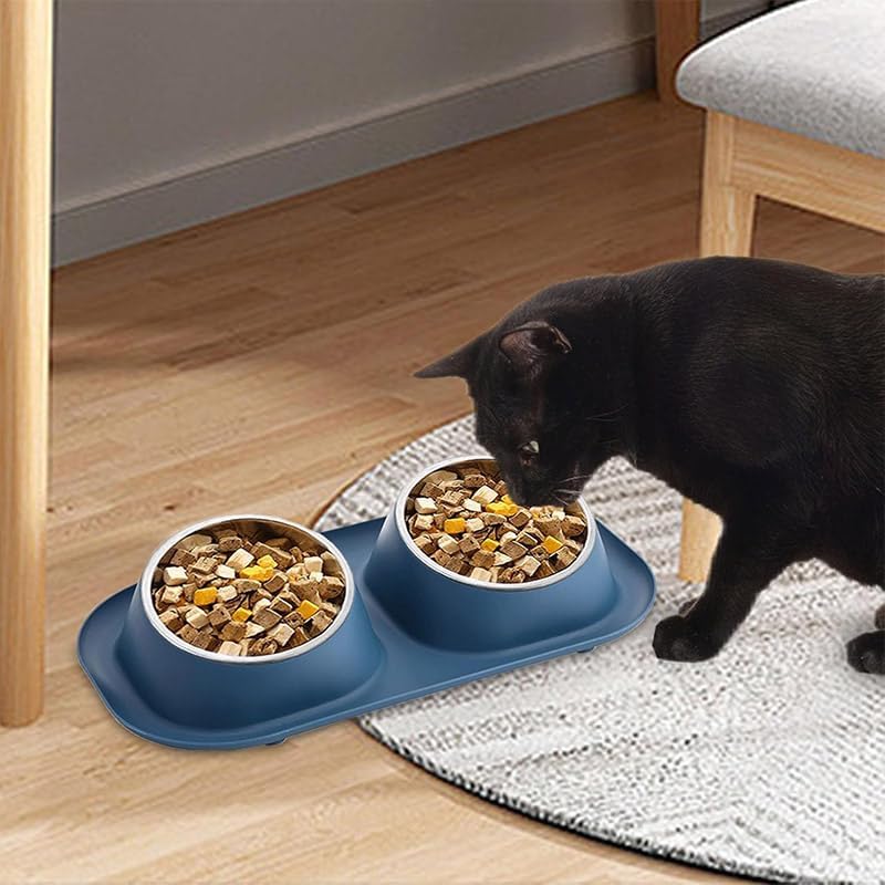 A black cat eating from two bowls of dry cat food placed on a blue dual-bowl pet feeder tray on a light wood floor next to a chair at Pet Servo