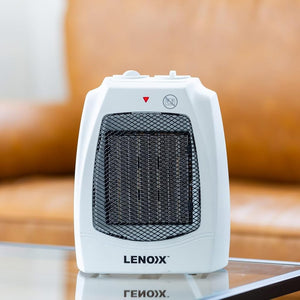 A white and black Lenox Gionex portable electric heater sits on a glass table with a blurred brown leather sofa in the background.