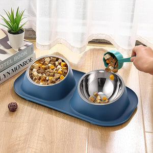 A blue dual pet food bowl set on a light wood floor, with one bowl filled with dry kibble and the other being filled with wet food from a teal scoop held by a human hand. A small potted plant and a book are in the background near a white curtain at Pet Servo
