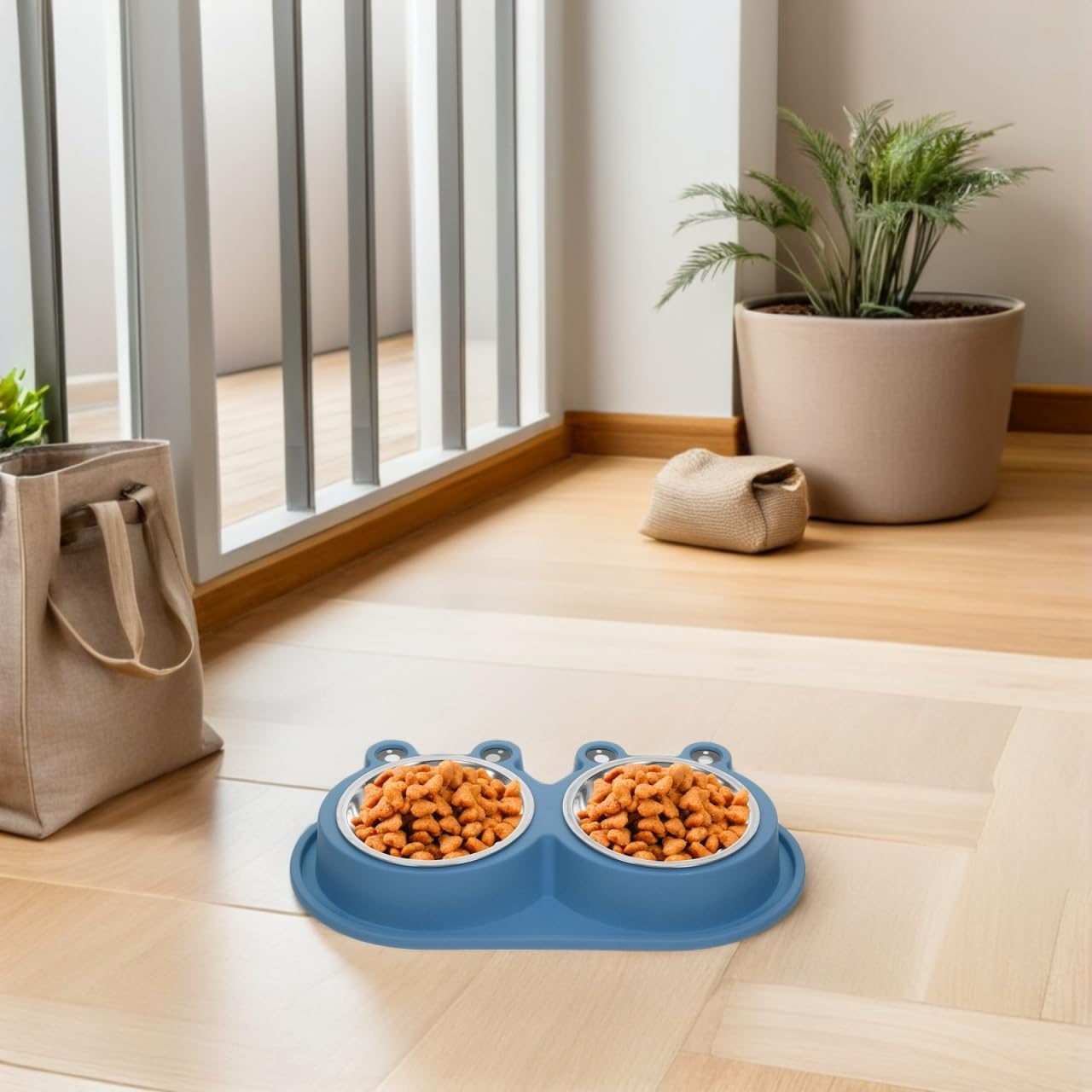 A blue dual pet food bowl with two handles sits on a light wood floor in a bright room, filled with dry dog food. A beige tote bag, a small pouch, and a potted plant are visible in the background near a window at Pet Servo
