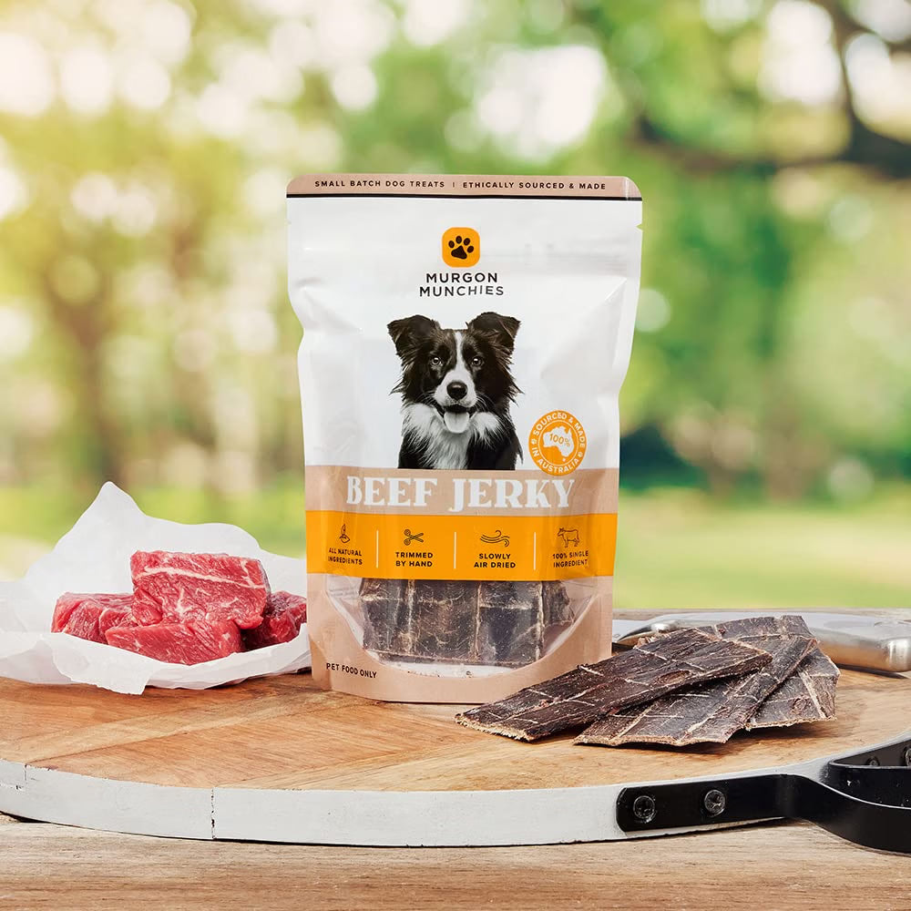 A bag of Murgon Munchies Beef Jerky dog treats stands on a wooden cutting board with raw meat pieces and sliced jerky pieces arranged around it. The background is a soft, blurred outdoor green garden setting.