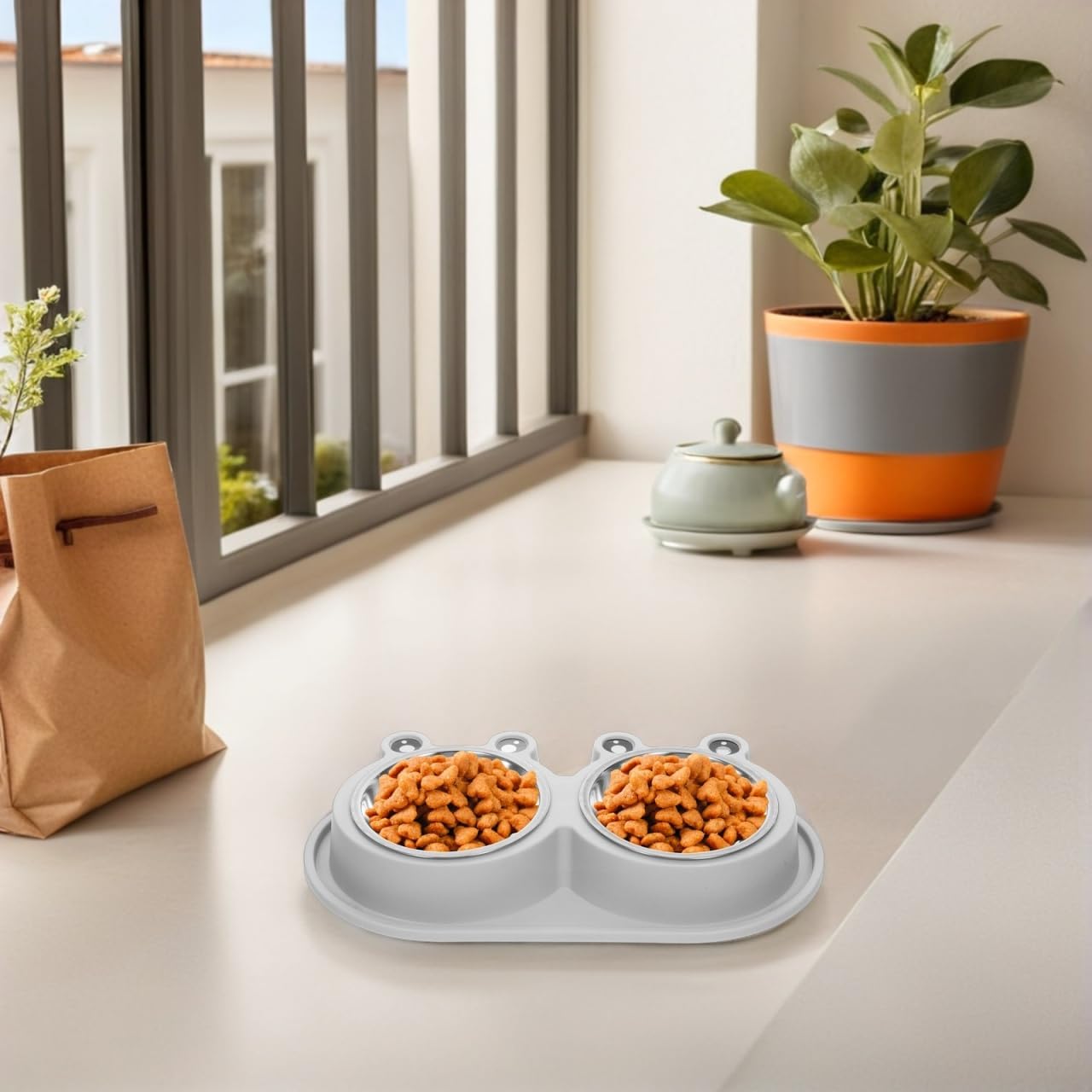 A dual pet food bowl set with a light gray silicone tray and two bowls filled with kibble, placed on a windowsill in a bright indoor setting with a plant and paper bag nearby at Pet Servo