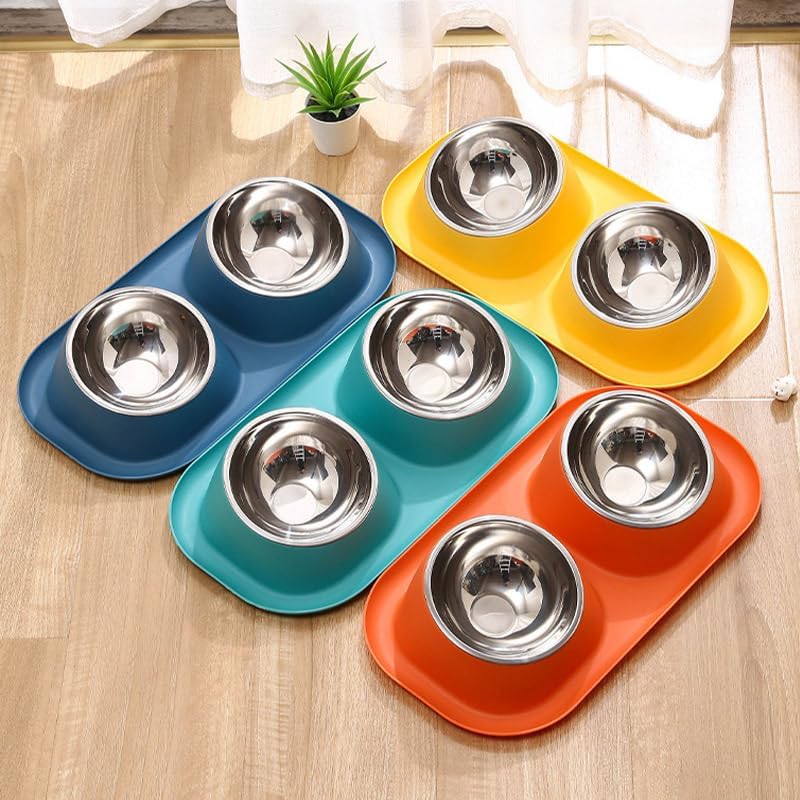 Four pet feeding trays in blue, teal, orange, and yellow, each with two stainless steel bowls, arranged on a light wood floor with a small potted plant in the background at Pet Servo