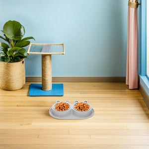 A gray dual pet food bowl filled with kibble sits on a light wood floor in a bright room with a blue wall, pink curtain, and a sisal cat tree with a plant at Pet Servo