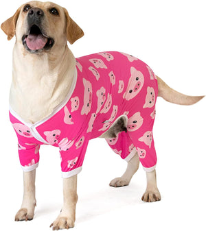 A light brown Labrador Retriever wearing a bright pink full-body dog pajama set with a white trim and a playful pink cartoon pig pattern, standing against a solid white background at Pet Servo