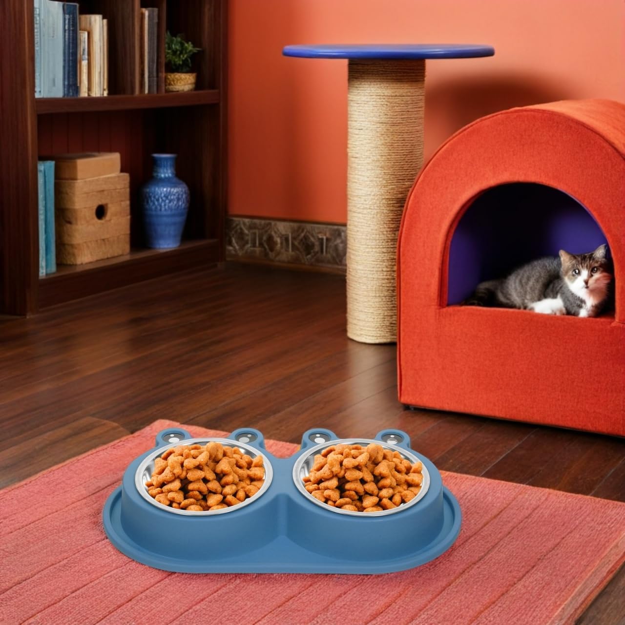A blue dual pet feeder bowl filled with dry cat food sits on a pink mat in a living room setting. A gray and white cat is visible inside an orange cat bed in the background, along with a scratching post and bookshelf at Pet Servo