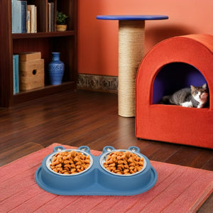 A blue dual pet feeder bowl filled with dry cat food sits on a pink mat in a living room setting. A gray and white cat is visible inside an orange cat bed in the background, along with a scratching post and bookshelf at Pet Servo