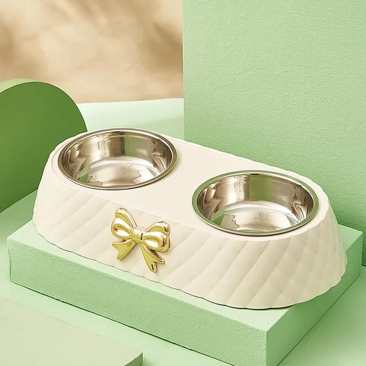 A double pet bowl set featuring a cream-colored ribbed plastic base with a shiny gold bow accent and two stainless steel bowls, displayed on a matching light green geometric pedestal at Pet Servo