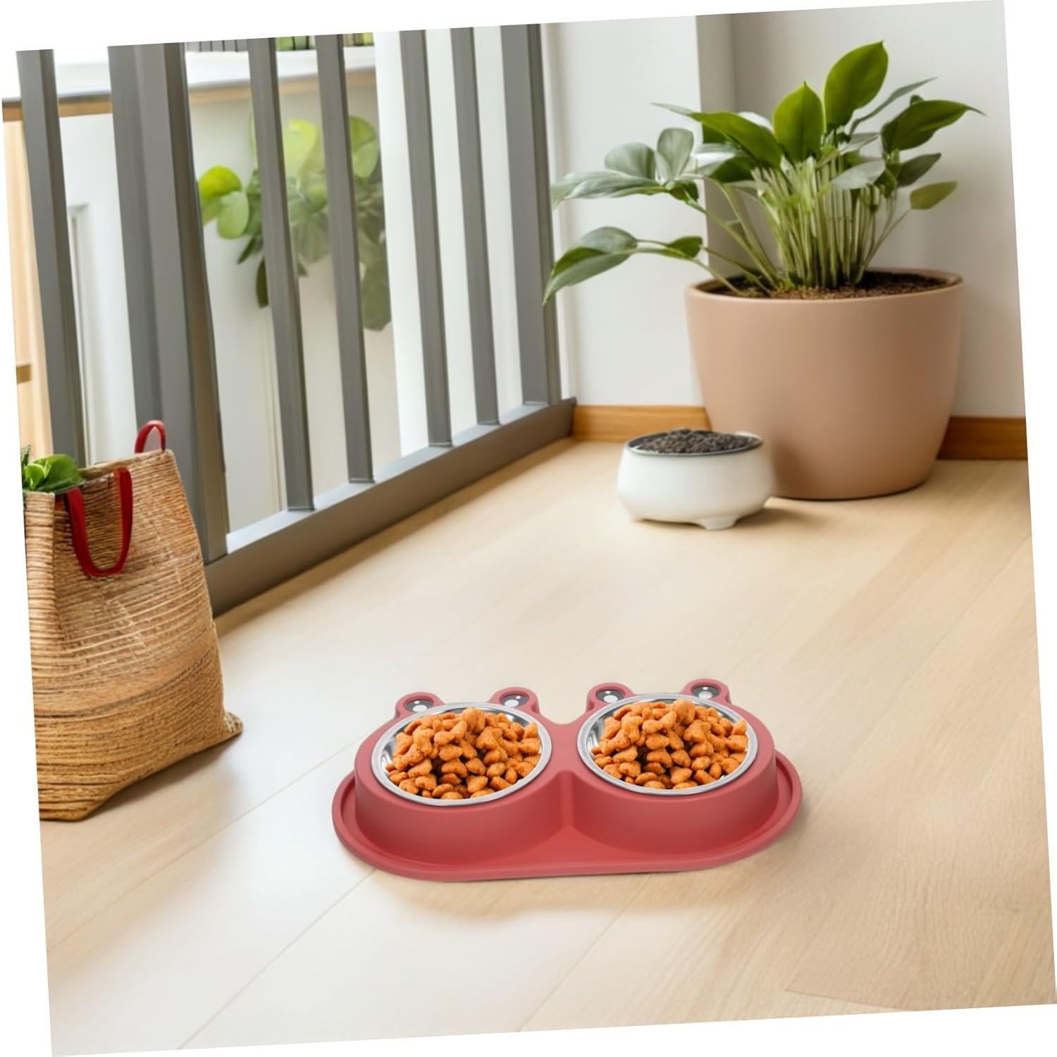 A pink dual-bowl pet feeder and waterer set on a light wood floor, with two stainless steel bowls filled with kibble. The scene is styled like a home interior with a potted plant, a straw bag, and a small white bowl in the background at Pet Servo