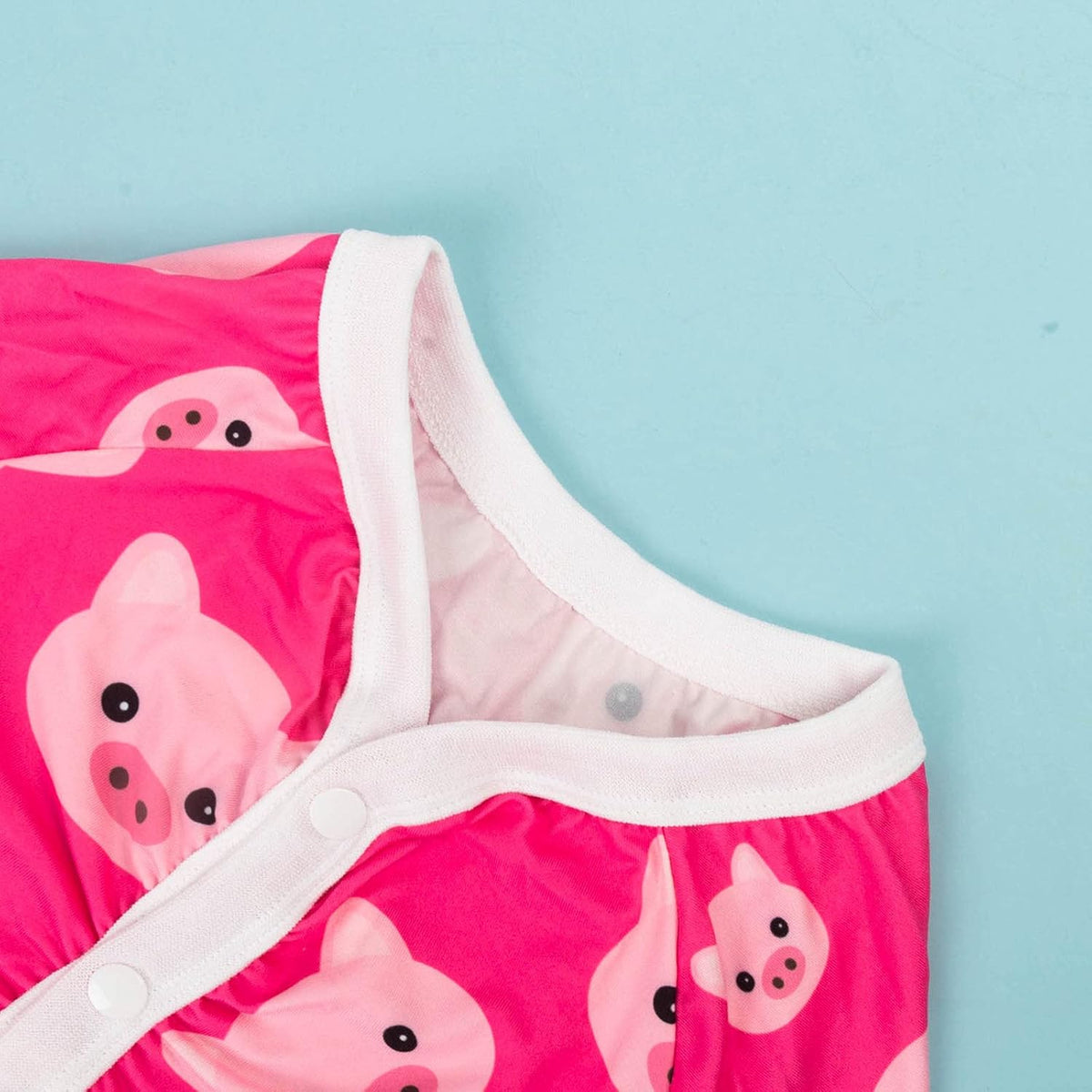 A close-up of a baby garment in bright pink with a repeating cartoon pig print and white trim, laid against a light blue background at Pet Servo