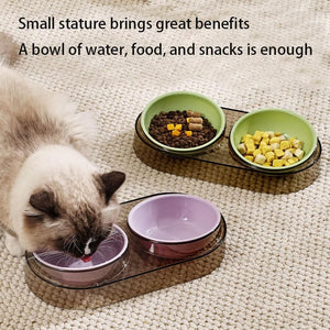 A cat with cream and dark brown fur is shown drinking water from one of two pet feeding trays on a textured beige rug. The trays hold bowls of food and snacks, including wet cat food, dry kibble, and yellow cubes at Pet Servo