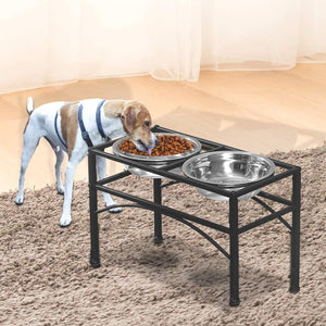 A small white and tan dog wearing a harness stands beside a black elevated double pet feeder with stainless steel bowls, one filled with kibble and one empty, on a beige shag rug in front of a light curtain at Pet Servo